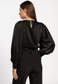 Zwarte satijnen blouse met opgezette mouwen en een sleutelgatdetail aan de achterkant, met een metallic rand rond de halslijn. Draagt bij zwarte, op maat gemaakte broeken.