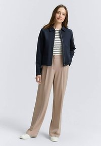 Veste zippée bleu marine avec un col, portée avec une chemise rayée et un pantalon large beige clair. Des chaussures blanches complètent l'ensemble.