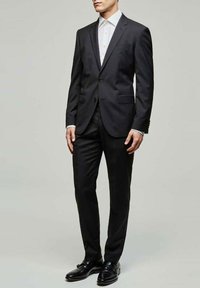 Costume noir ajusté avec une veste à un seul bouton, un pantalon assorti et une chemise blanche. Terminé avec des chaussures en cuir noires.