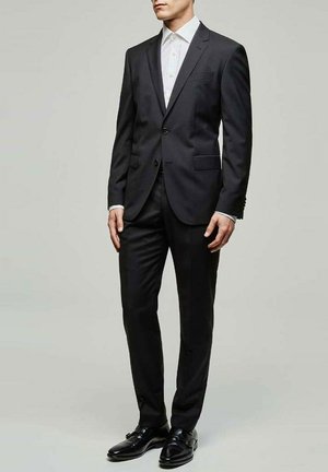 Costume noir ajusté avec une veste à un seul bouton, un pantalon assorti et une chemise blanche. Terminé avec des chaussures en cuir noires.