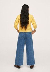 Pull jaune associé à un jean bleu à jambes larges. Le pull possède des poignets côtelés ; le jean a des poches plaquées et des ourlets effilochés.