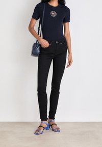 Top de manga corta azul marino con logo redondo, jeans ajustados negros, sandalias de tacón bloque azul y bolso cruzado pequeño azul.