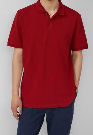 Homme portant un polo rouge uni à manches courtes et un pantalon bleu marine, se tenant devant un fond gris clair.