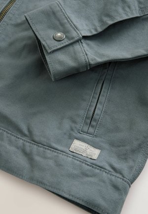 Jachetă de culoare albastru deschis, realizată dintr-un material texturat, cu închidere prin fermoar și un buton tip clapetă pe manșetă. Include un etichete cu marcaj pe tiv.