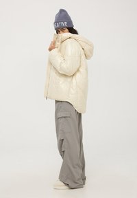 Kremowa kurtka puchowa z kapturem, wyróżniająca się pikowaną teksturą, zestawiona z luźnymi szarymi spodniami cargo i szarym beanie z napisem "CREATIVE".