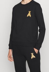 Felpa nera con polsini e orlo a costine, presenta un design dorato di un insetto sul petto. Tessuto comodo e elegante, adatto per un abbigliamento casual.