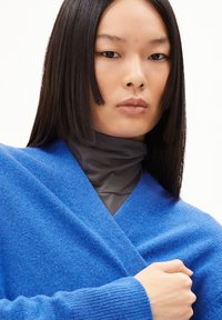 Levendig blauwe wollen cardigan met een V-halsontwerp, gestructureerde stof en geribde manchetten, gelaagd over een grijze coltrui.