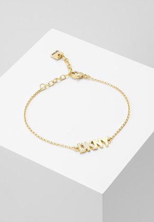 Bracelet chaîne en or avec fermoir et maillons ajustables, arborant le logo "DKNY" orné de petits cristaux clairs, présenté sur une surface blanche.