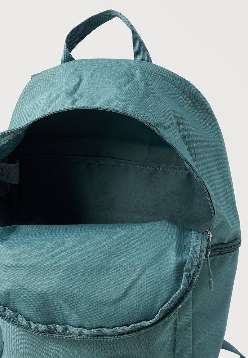 Mochila de tela color verde azulado con el compartimento principal abierto que muestra el interior vacío y una asa superior para transportar, sobre un fondo blanco.