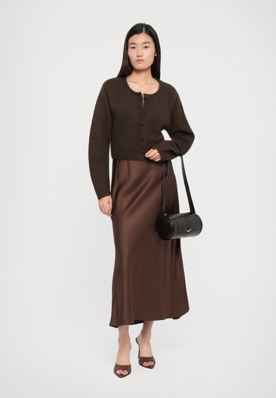 Brown cropped cardigan with buttons, worn over a brown satin maxi dress. Accessories include a black, round handbag with a strap.