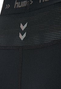 Black athletic leggings with a textured waistband, featuring a mesh area and reflective chevron design. Smooth fabric with detailed stitching.