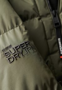Chaqueta acolchada verde oliva con un sutil brillo, que presenta el logo bordado en negro "SUPERDRY JPN" en la manga y detalles de herrajes en contraste.