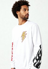 Man met krullen, gekleed in een wit shirt met lange mouwen, met een gele bliksemschicht op de borst, zwarte vlammen en rode tekst op de mouwen.