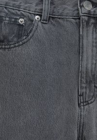 Gros plan sur un jean denim gris montrant la poche avant, la passoire de ceinture et les rivets métalliques avec estampillage de la marque.