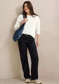 Pull en tricot blanc avec un motif texturé, associé à un jean foncé et des baskets de couleur claire. Un sac en tissu bleu pend sur une épaule.