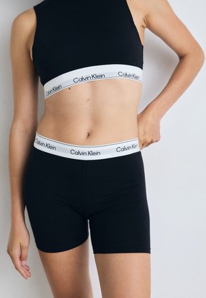 Black stretch fabric bralette and shorts with a white Calvin Klein logo band at the waist. Close-fitting, smooth texture, casual design.