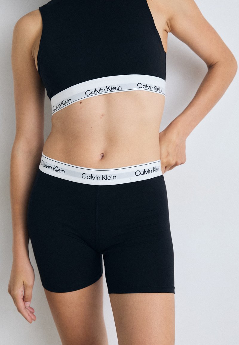 Bralette e pantaloni corti in tessuto elasticizzato nero con una fascia con logo Calvin Klein bianco in vita. Vestibilità aderente, superficie liscia, design casual.