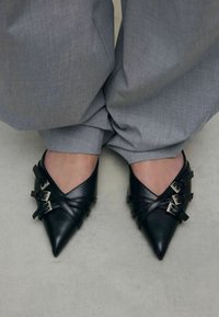 Chaussures en cuir noir à bout pointu avec des sangles croisées et des boucles en argent. Portées avec un pantalon plissé gris clair. Texture lisse.