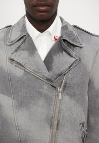 Jachetă de motociclist din denim gri cu o textură uzată, fermoar asimetric și rever pronunțat. Are o cămașă albă pe dedesubt.