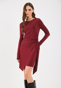 Vestido bordô de mangas longas com detalhe lateral torcido e bainha assimétrica. Feito de um tecido liso e elástico. Botas pretas até o joelho.