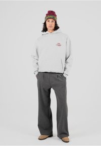 Sudadera gris con bolsillo frontal, logo de marca en burdeos; combinada con pantalones anchos grises y zapatos beige. Gorro de punto burdeos con acentos en verde.