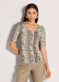 Vrouw in een dierenprint t-shirt met korte mouwen en beige broek, staand met één hand op de heup tegen een effen achtergrond.