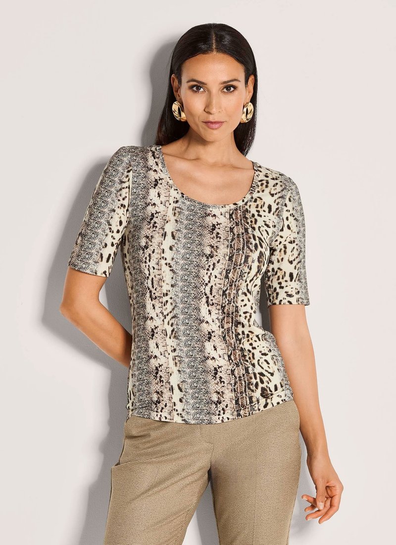 Vrouw in een dierenprint t-shirt met korte mouwen en beige broek, staand met één hand op de heup tegen een effen achtergrond.