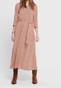 Pitkä, vaaleanlila mekko, jossa on kukkakuosi, nappikiinnitys edessä, kolmivartiset hihat, sidottu vyötärö ja valuva helma. Päällä ruskeat nilkkurit.