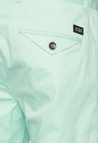 Jasnozielone szorty w kolorze mięty z tylną kieszenią w kształcie rombu, zapinaną na guzik, oraz małą czarną metką marki wszytą po boku.