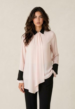 Donna con capelli lunghi e scuri che indossa una camicetta rosa pallido con colletto e polsini neri, abbinata a pantaloni neri, in piedi davanti a uno sfondo semplice.