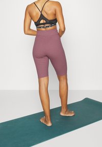 Reggiseno sportivo nero con design a taglio e spalline regolabili, abbinato a pantaloni da bicicletta a coste color malva, mostrato da dietro su un tappetino da yoga.