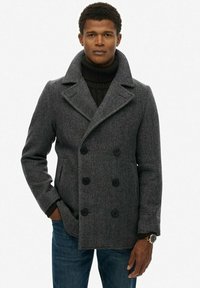 Abrigo de lana gris con un diseño de doble botonadura, cuello ancho y grandes botones negros. Lleva puesto sobre un cuello alto negro, combinado con jeans azules.