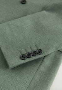 Manica di un abito verde con quattro bottoni neri in fila vicino al polsino, che mostra i dettagli della fine tessitura del tessuto.