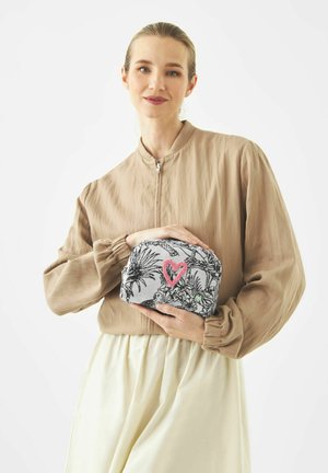 Frau trägt eine beige Jacke und einen cremefarbenen Rock und hält eine graue Tasche mit schwarzem Blumenmuster und einem pinken Herzdesign.