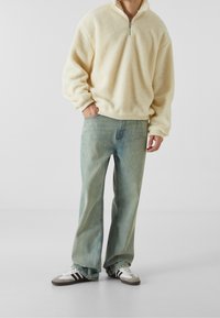 Plyšový pulovr v jemné žluté barvě s čtvrtinovým zipem, spárovaný se světle modrými rovnými džíny a bílými teniskami se černými pruhy.