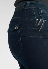 Jeans de mezclilla azul oscuro con un diseño ajustado, que cuentan con un pequeño bolsillo con cremallera, una textura sutil y costuras de contraste en los bolsillos traseros.