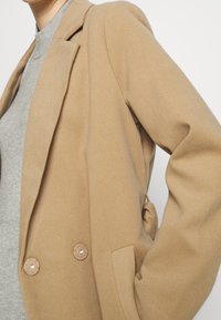 Osoba nosząca beżowy wełniany płaszcz na jasnoszarym swetrze, z ręką w kieszeni płaszcza na jednolitym białym tle.