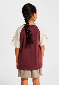 Vinröd kortärmad tröja med beiga raglanärmar och tre beige ränder på vänster ärm. Bärs med beiga shorts, slät textur.