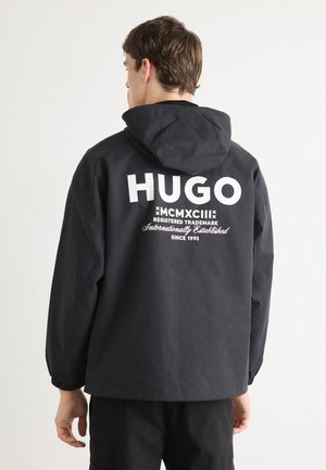 Veste à capuche noire avec un texte logo blanc au dos. Dotée de poignets élastiques et d'une coupe décontractée. Fabriquée en matériau léger et résistant à l'eau.