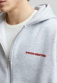 Sweat à zip gris en tissu doux, avec le mot "INTERDIT" brodé en rouge sur la poitrine et une grande capuche. En dessous, il y a une chemise blanche.
