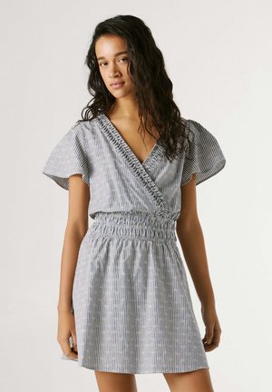 Giovane donna con capelli lunghi e scuri che indossa un vestito corto a righe con scollo a V, maniche svolazzanti e vita arricciata, in piedi davanti a uno sfondo semplice.