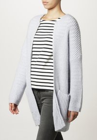 Hellgrauer, gestrickter Cardigan mit geripptem Muster, offener Vorderseite und zwei vorderen Taschen, getragen über einem gestreiften Langarmshirt.