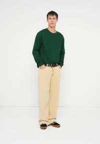 Groene longsleeve top, crèmekleurige wijde broek, zwarte riem met gouden accenten en zwarte instapschoenen met witte details.