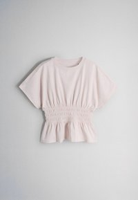 Hele roosa lühikeste varrukatega pluus, millel on kohapeal kogutud elastne smockitud vöökoht ja kergelt lõhestatud peplum-äär, riputatud lihtsa valge tausta vastu.