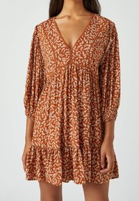 Donna che indossa un vestito floreale arancione ruggine con scollatura a V profonda, maniche a 3/4 a sbuffo e orlo arricciato su uno sfondo semplice.