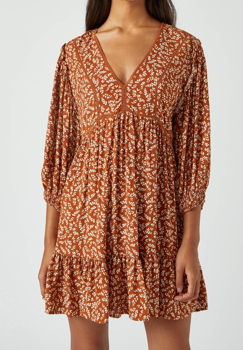 Donna che indossa un vestito floreale arancione ruggine con scollatura a V profonda, maniche a 3/4 a sbuffo e orlo arricciato su uno sfondo semplice.