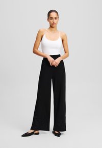 Zwarte wijde broek met bloemembroidery aan de zoom, gecombineerd met een witte aansluitende tanktop. Model toont een neutrale blik.