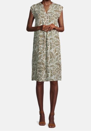 Kvinde iført knælængde, ærmeløs kjole med grønne og hvide bladmønstre og brune ankel-rem hælede sandaler, stående med hænderne foldet.