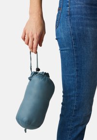 Kompakte türkisfarbene Tasche aus glattem Material, oben gerafft, von einer Hand mit Kordelzug und Stopper gehalten, neben einem in Jeans gekleideten Bein.