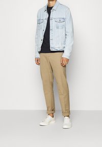 Giacca di jeans azzurro chiaro con due tasche sul petto, camicia nera sotto, pantaloni khaki a gamba affusolata e sneakers bianche, in piedi su uno sfondo semplice.